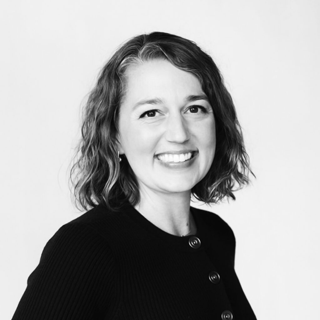 Dr. Ashley Nelson, a DC (Doctor of Chiropractic), AFMCP (Applying Functional Medicine in a Clinical Practice) in a studio portrait against a light gray background wearing a black sweater with gold buttons.