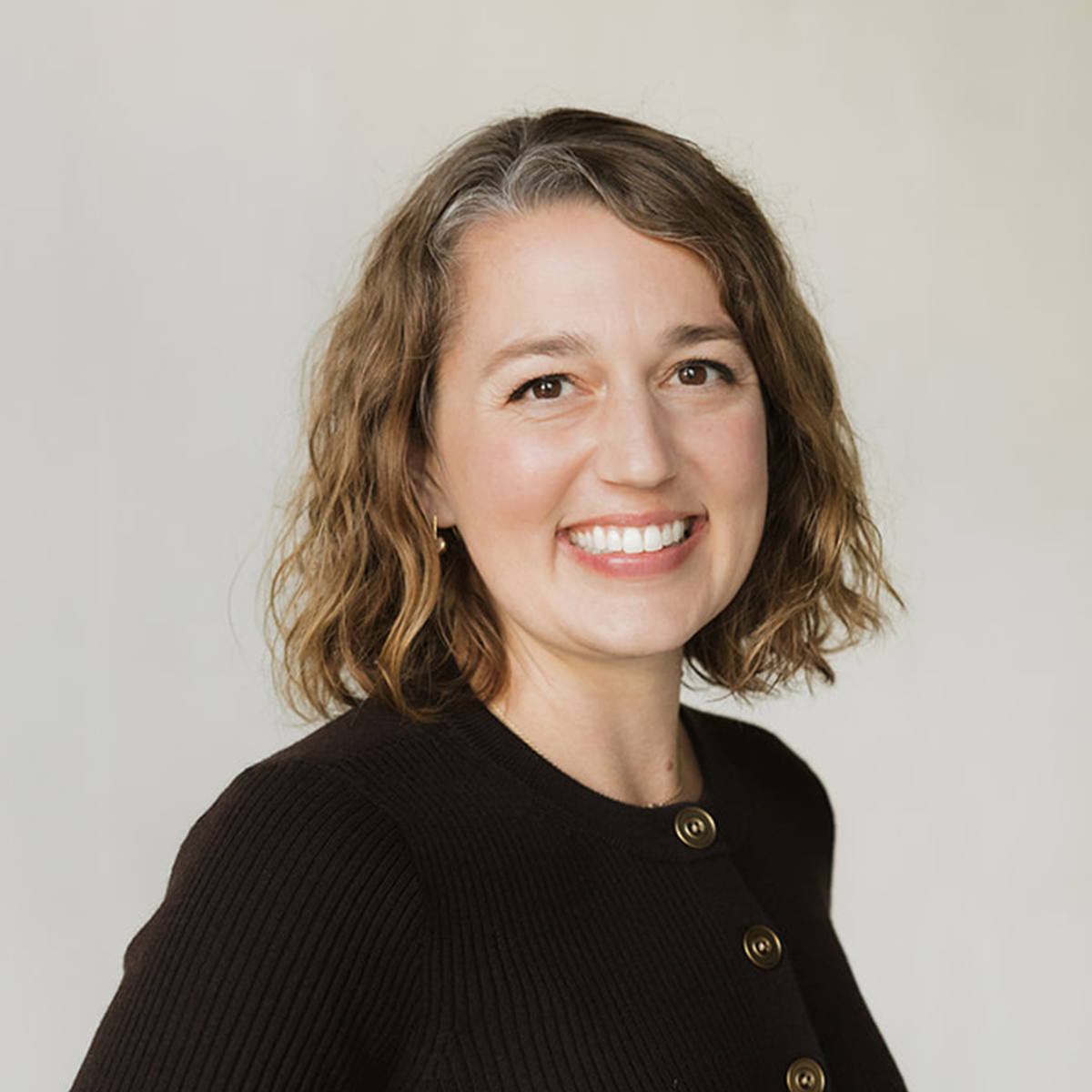Dr. Ashley Nelson, a DC (Doctor of Chiropractic), AFMCP (Applying Functional Medicine in a Clinical Practice) in a studio portrait against a light gray background, wearing a black sweater with gold buttons.