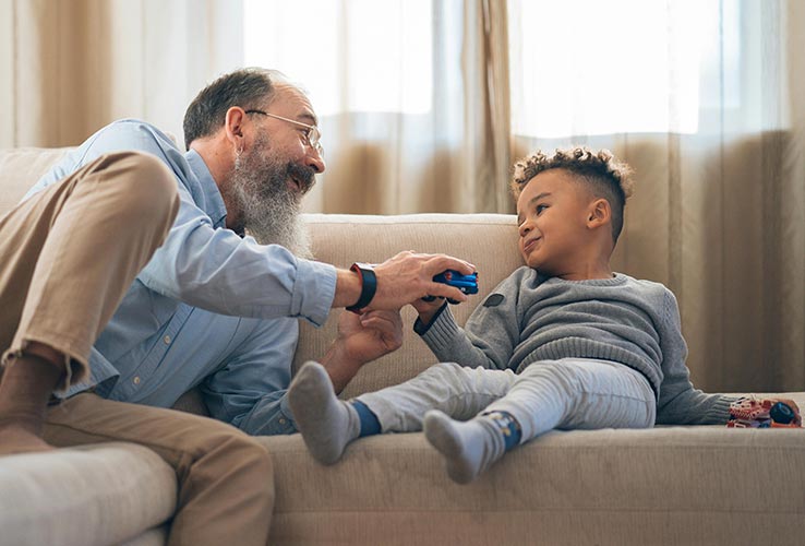 An older man and a young boy sit on a couch, smiling as they play with toy cars in a living room with soft natural light.