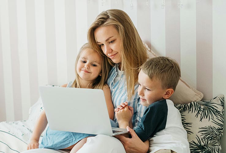 A woman sits on a bed with two young children, all looking at a laptop together in a bright room with striped walls and patterned pillows.