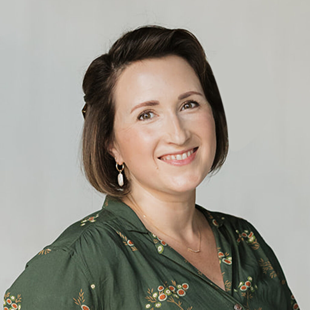 Becki O’Brien in a studio portrait against a light gray background, wearing a dark green floral blouse and gold earrings.