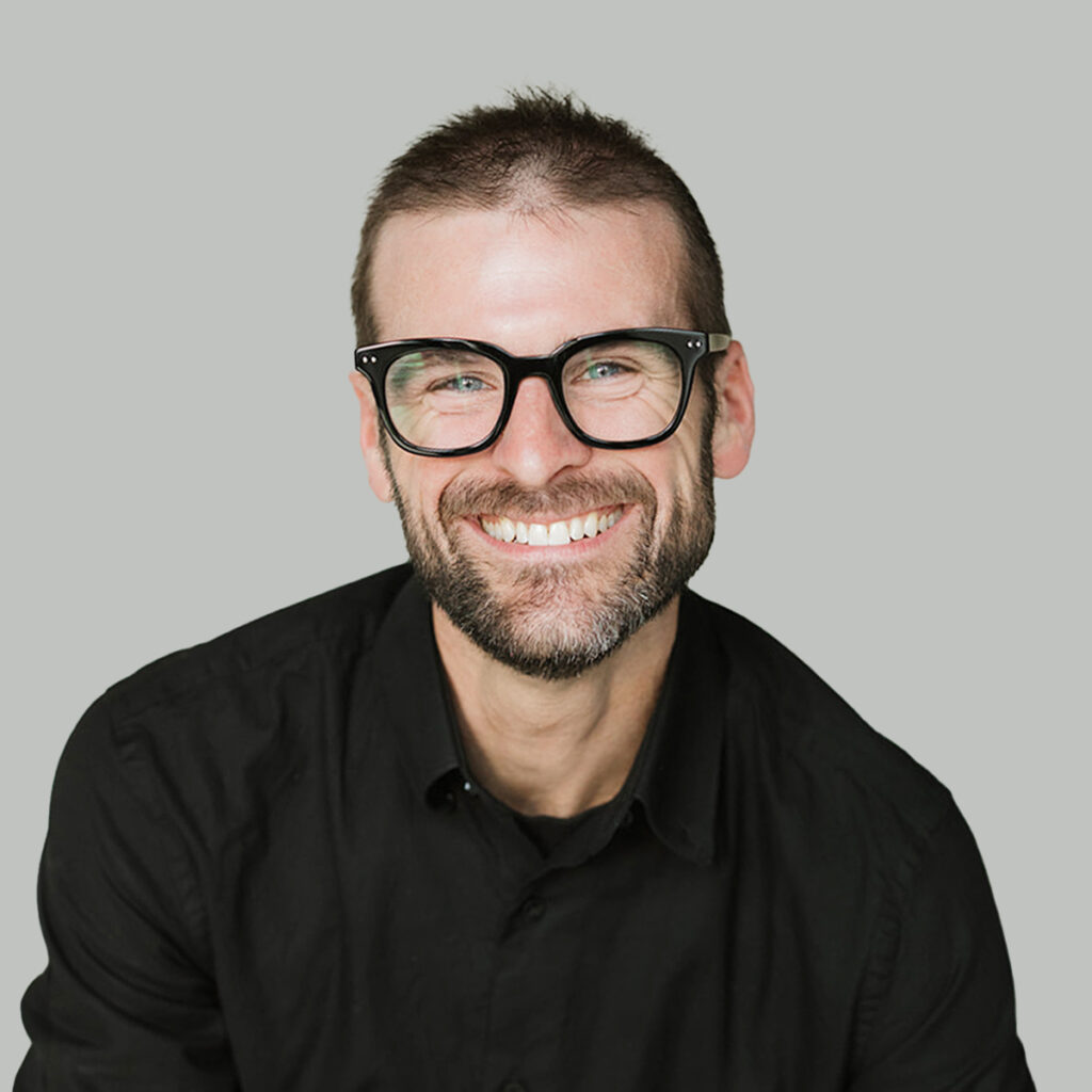 Tim O’Brien smiles in a studio portrait against a light gray background, wearing black glasses and a black button-up shirt.